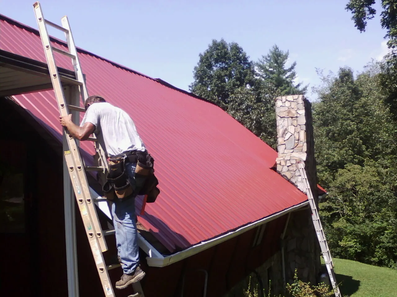 Roofer on a ladder beside a red metal roof and stone chimney — Soffit & Fascia Repair by Roofing Brothers in New Market